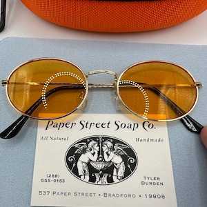 May include: Round, amber-tinted sunglasses with a gold metal frame. The sunglasses are resting on a business card that reads "Paper Street Soap Co. All Natural Handmade (288) 555-0153 TYLER DURDEN 537 PAPER STREET BRADFORD 19808".
