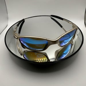 May include: Silver-framed sunglasses with blue-tinted lenses rest on a reflective surface. The sunglasses have a sleek, modern design with a curved frame and black accents on the temples. The lenses reflect a spectrum of colors.