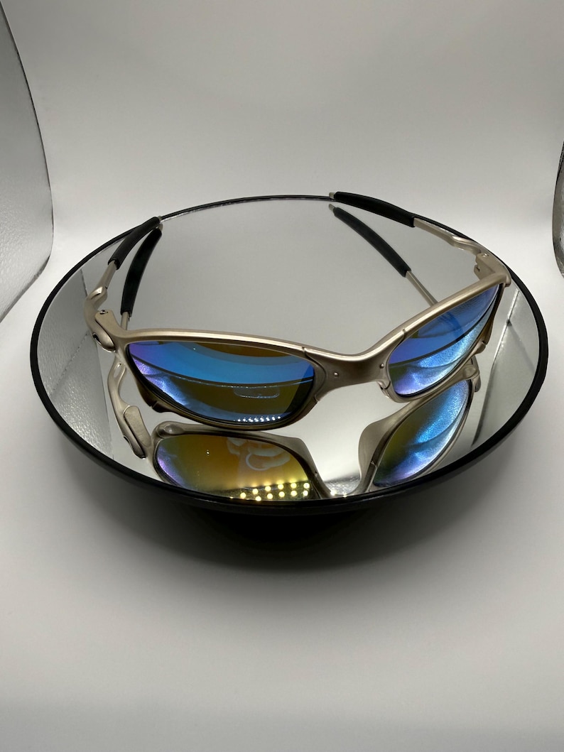 May include: Silver-framed sunglasses with blue-tinted lenses rest on a reflective surface. The sunglasses have a sleek, modern design with a curved frame and black accents on the temples. The lenses reflect a spectrum of colors.