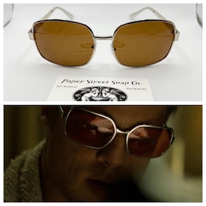 May include: Silver-framed sunglasses with brown lenses. The sunglasses have a rectangular shape and are resting on a white surface. Below the sunglasses is a white card with the text "Paper Street Soap Co."