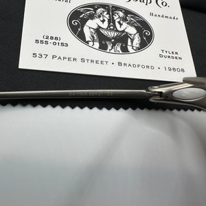 May include: A business card for Paper Street Soap Co. is displayed, featuring a black and white illustration of cherubs. The card includes the text "All Natural" and "Handmade", along with a phone number and address. A silver tool is partially visible.