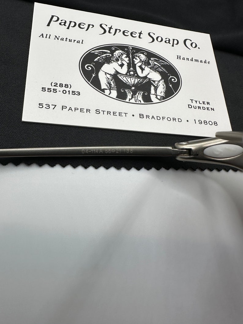 May include: A business card for Paper Street Soap Co. is displayed, featuring a black and white illustration of cherubs. The card includes the text "All Natural" and "Handmade", along with a phone number and address. A silver tool is partially visible.