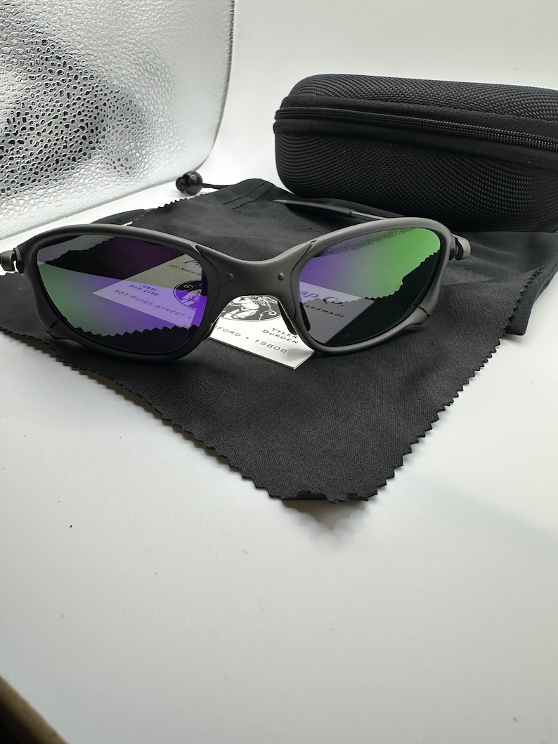 May include: A pair of black sunglasses with green and purple mirrored lenses. The sunglasses are on a black microfiber cloth with a small white tag that reads "Tyler Duncan Handmade, 82 Paper Street, Ford, 19808".