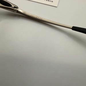Peut inclure: Gros plan sur une monture de lunettes argent&eacute;e avec le texte "JULIET CE" grav&eacute; sur la branche. La monture a un design &eacute;l&eacute;gant et moderne avec des accents noirs sur les extr&eacute;mit&eacute;s des branches. Une carte blanche avec du texte noir est en arri&egrave;re-plan.