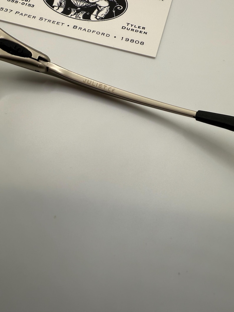 Peut inclure: Gros plan sur une monture de lunettes argent&eacute;e avec le texte "JULIET CE" grav&eacute; sur la branche. La monture a un design &eacute;l&eacute;gant et moderne avec des accents noirs sur les extr&eacute;mit&eacute;s des branches. Une carte blanche avec du texte noir est en arri&egrave;re-plan.