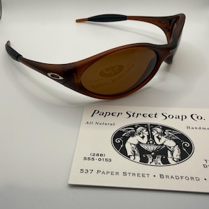 Puede incluir: Gafas de sol Oakley marrones con montura curvada y lentes tintadas. Las gafas de sol tienen detalles negros en las patillas y el logotipo de Oakley es visible en la montura. Una tarjeta de visita de Paper Street Soap Co. está en primer plano.