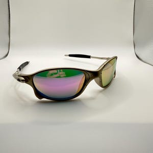 May include: A pair of metallic sunglasses with a curved frame and iridescent lenses. The lenses reflect a spectrum of colors, including pink, green, and yellow. The temples have black tips.