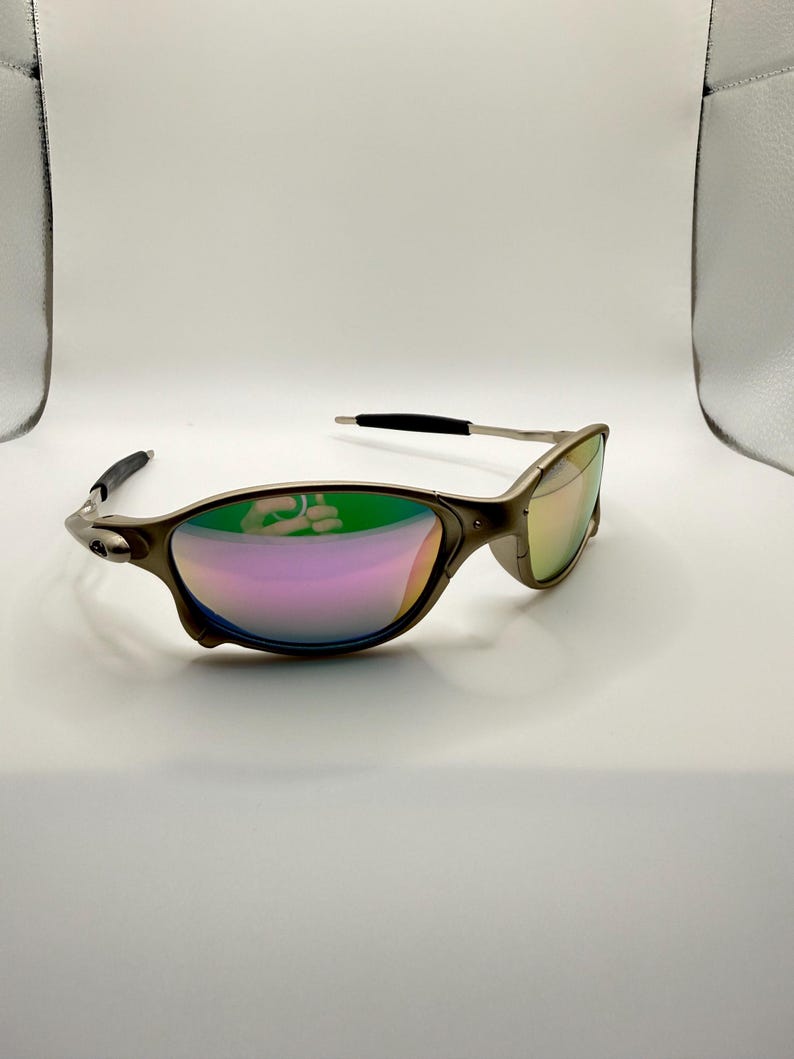 May include: A pair of metallic sunglasses with a curved frame and iridescent lenses. The lenses reflect a spectrum of colors, including pink, green, and yellow. The temples have black tips.