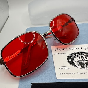 May include: Red square-shaped sunglasses with a silver metal frame. The sunglasses are on a blue surface with a black cleaning cloth and a business card that reads "Paper Street Soaps, All Natural (288) 555-0153 537 Paper Street".