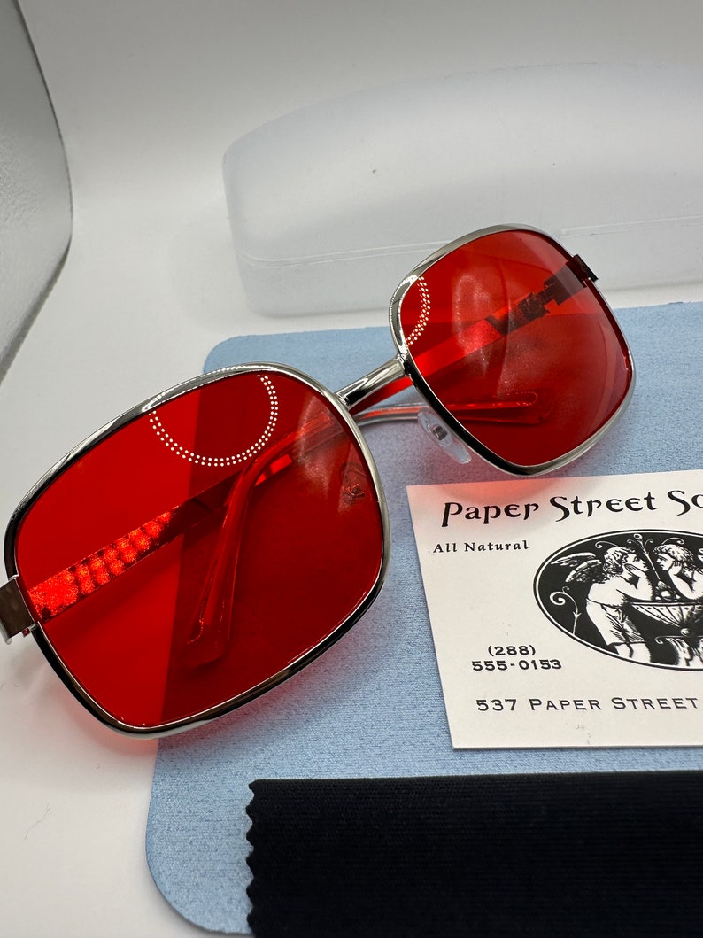 May include: Red square-shaped sunglasses with a silver metal frame. The sunglasses are on a blue surface with a black cleaning cloth and a business card that reads "Paper Street Soaps, All Natural (288) 555-0153 537 Paper Street".