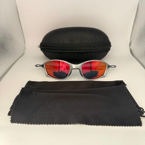 May include: Silver-framed sunglasses with red lenses, a black zippered case, and a black microfiber cleaning cloth. The sunglasses have a sleek, modern design. The case is oval-shaped. The cloth is folded.