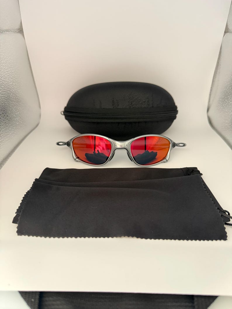 May include: Silver-framed sunglasses with red lenses, a black zippered case, and a black microfiber cleaning cloth. The sunglasses have a sleek, modern design. The case is oval-shaped. The cloth is folded.