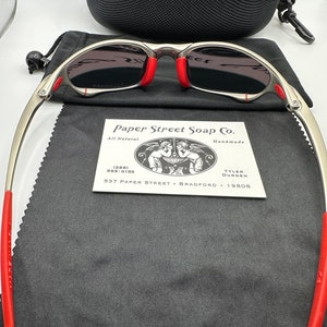 K&ouml;nnte beinhalten: Silberfarbene Sonnenbrille mit roten Akzenten und grauen Gl&auml;sern. Die Sonnenbrille liegt auf einem schwarzen Mikrofaser-Reinigungstuch. Eine kleine Visitenkarte mit dem Text "Paper Street Soap Co. All Natural Handmade (288) 555-0153 TYLER DURDEN 537 PAPER STREET BRADFORD 19808" ist im Bild zu sehen.