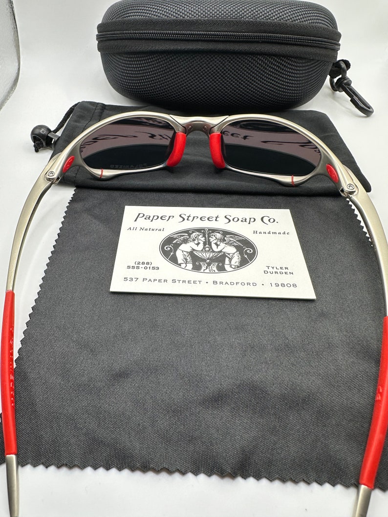 K&ouml;nnte beinhalten: Silberfarbene Sonnenbrille mit roten Akzenten und grauen Gl&auml;sern. Die Sonnenbrille liegt auf einem schwarzen Mikrofaser-Reinigungstuch. Eine kleine Visitenkarte mit dem Text "Paper Street Soap Co. All Natural Handmade (288) 555-0153 TYLER DURDEN 537 PAPER STREET BRADFORD 19808" ist im Bild zu sehen.