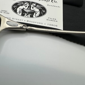 May include: A close-up of a pair of silver-toned scissors with the text "JULIET CE" engraved on the blade. A business card for Paper Street Soap Co. is in the background, with a black cloth. The card features an illustration of two angels.
