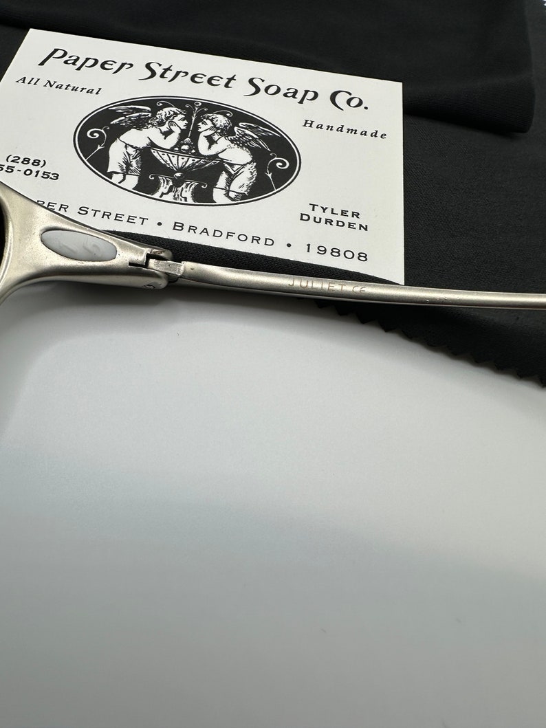 May include: A close-up of a pair of silver-toned scissors with the text "JULIET CE" engraved on the blade. A business card for Paper Street Soap Co. is in the background, with a black cloth. The card features an illustration of two angels.