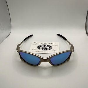 May include: Sunglasses with a metallic frame and blue lenses. The sunglasses have black arms and a small white card with the text "Paper Street Soap Co." is behind the glasses.
