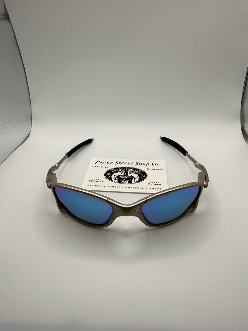 May include: Sunglasses with a metallic frame and blue lenses. The sunglasses have black arms and a small white card with the text "Paper Street Soap Co." is behind the glasses.