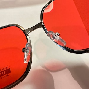 Puede incluir: Primer plano de unas gafas de sol cuadradas rojas con montura plateada. Las gafas de sol tienen almohadillas nasales de pl&aacute;stico transparente y el texto "Against UV 100% E" es visible en la lente izquierda.