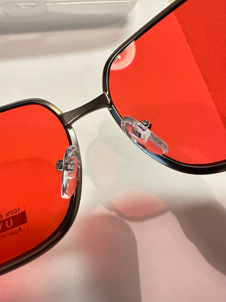 Puede incluir: Primer plano de unas gafas de sol cuadradas rojas con montura plateada. Las gafas de sol tienen almohadillas nasales de pl&aacute;stico transparente y el texto "Against UV 100% E" es visible en la lente izquierda.