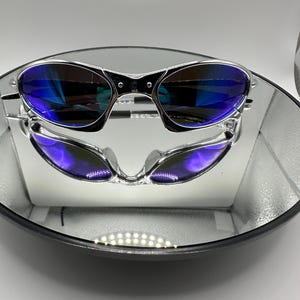 Op de afbeelding: Zilverkleurige zonnebril met blauwe glazen weerspiegeld in een ronde spiegel. De zonnebril heeft een strak, modern design. De glazen hebben een gradi&euml;nteffect, van blauw naar paars.