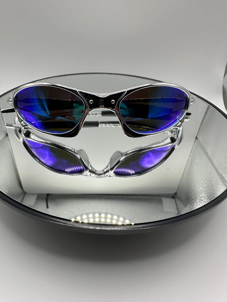 Op de afbeelding: Zilverkleurige zonnebril met blauwe glazen weerspiegeld in een ronde spiegel. De zonnebril heeft een strak, modern design. De glazen hebben een gradi&euml;nteffect, van blauw naar paars.