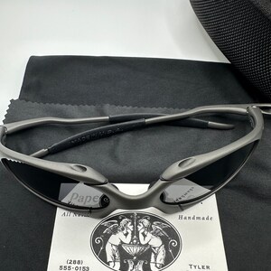 May include: A pair of gray metal sunglasses with a black case and a cleaning cloth. The sunglasses have a unique design with a curved frame and lenses. The case is made of a soft, black material and has a zipper closure. The cleaning cloth is made of a soft, gray material.