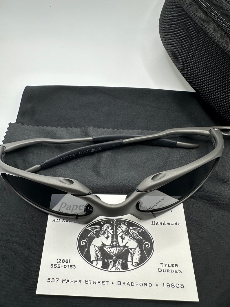 May include: A pair of gray metal sunglasses with a black case and a cleaning cloth. The sunglasses have a unique design with a curved frame and lenses. The case is made of a soft, black material and has a zipper closure. The cleaning cloth is made of a soft, gray material.