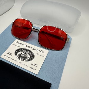 May include: Red square-shaped sunglasses with a silver metal frame. The sunglasses are on a blue suede surface with a black microfiber cleaning cloth. A white plastic case and a business card for Paper Street Soap Co. are also visible.