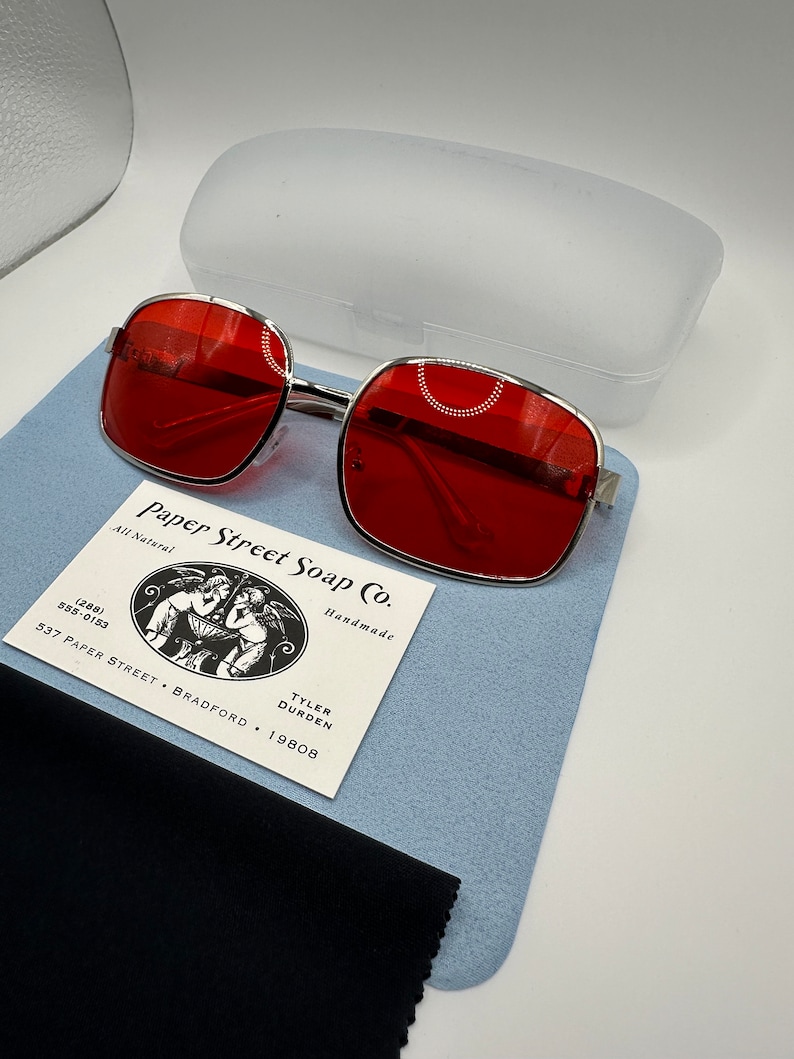May include: Red square-shaped sunglasses with a silver metal frame. The sunglasses are on a blue suede surface with a black microfiber cleaning cloth. A white plastic case and a business card for Paper Street Soap Co. are also visible.