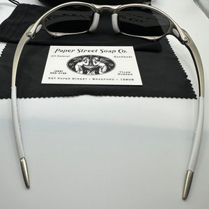 May include: Silver-framed sunglasses with dark lenses, white nose pads, and white temple tips. The glasses rest on a black cloth, with a black case and a business card from Paper Street Soap Co. visible in the background.