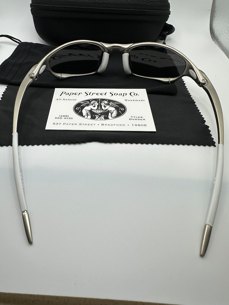 May include: Silver-framed sunglasses with dark lenses, white nose pads, and white temple tips. The glasses rest on a black cloth, with a black case and a business card from Paper Street Soap Co. visible in the background.