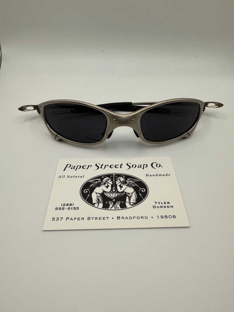 Peut inclure: Des lunettes de soleil argent&eacute;es &agrave; verres fonc&eacute;s sont pos&eacute;es sur une carte de visite. La carte indique "Paper Street Soap Co." avec une illustration et des coordonn&eacute;es, dont une adresse.