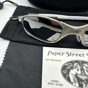 May include: Silver-framed sunglasses with reflective lenses, resting on a black cloth. The glasses are next to a black case and a tag that reads "Paper Street" with a phone number. The glasses have a white arm and are marked "MADE IN IT".