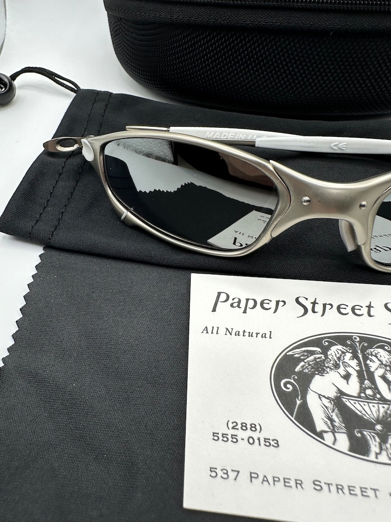 May include: Silver-framed sunglasses with reflective lenses, resting on a black cloth. The glasses are next to a black case and a tag that reads "Paper Street" with a phone number. The glasses have a white arm and are marked "MADE IN IT".