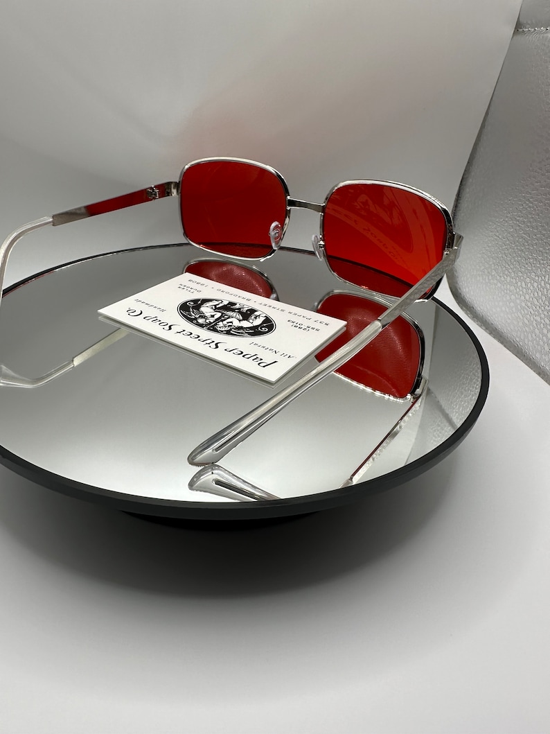 May include: Silver framed sunglasses with red lenses. The sunglasses are sitting on a mirrored surface with a business card for "Paper Street Soap Co." in the background.