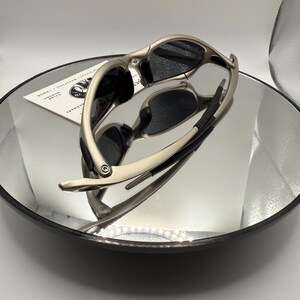 Peut inclure: Des lunettes de soleil argent&eacute;es avec des verres noirs sont pr&eacute;sent&eacute;es sur une surface r&eacute;fl&eacute;chissante. Les lunettes de soleil ont un design &eacute;l&eacute;gant et moderne avec une monture incurv&eacute;e et des verres fonc&eacute;s. L'image est bien &eacute;clair&eacute;e, soulignant la finition m&eacute;tallique des montures.
