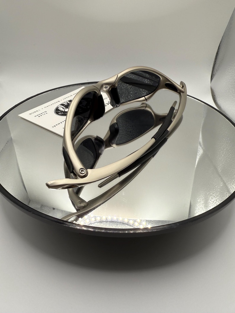 Peut inclure: Des lunettes de soleil argent&eacute;es avec des verres noirs sont pr&eacute;sent&eacute;es sur une surface r&eacute;fl&eacute;chissante. Les lunettes de soleil ont un design &eacute;l&eacute;gant et moderne avec une monture incurv&eacute;e et des verres fonc&eacute;s. L'image est bien &eacute;clair&eacute;e, soulignant la finition m&eacute;tallique des montures.