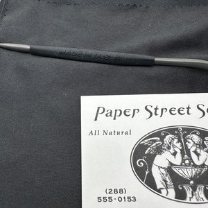 May include: A white business card with black text that reads "Paper Street So", "All Natural", "(288)", "555-0153", and "537 PAPER STREET E". The card features a black and white illustration of two angels.