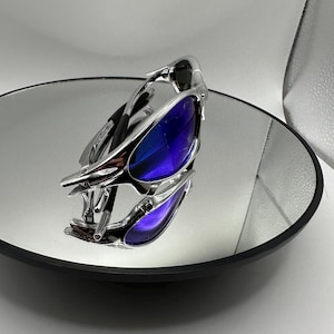 Op de afbeelding: Een futuristische zonnebril met een zilverkleurig montuur en blauw-paarse glazen. De bril ligt op een ronde spiegel, die het ontwerp weerspiegelt. De glazen hebben een kleureffect van blauw naar paars.