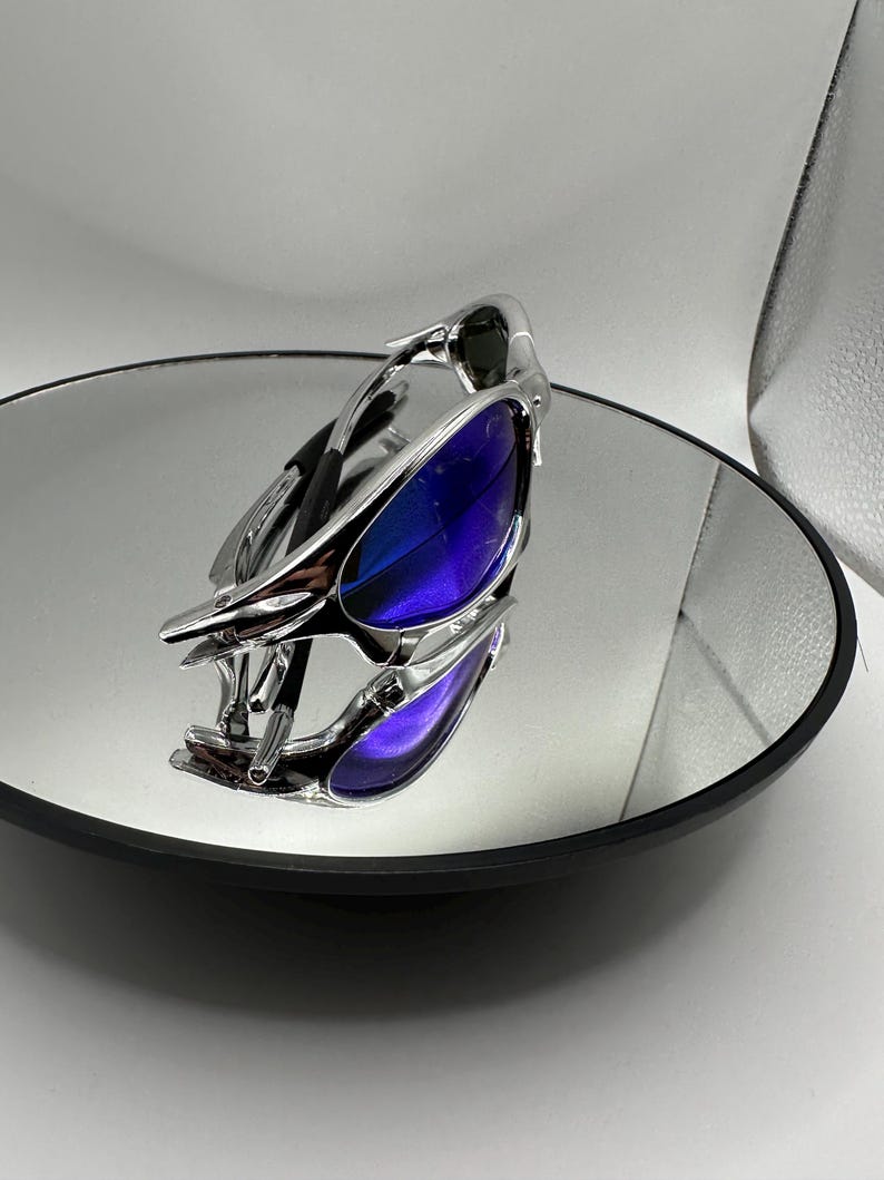 Op de afbeelding: Een futuristische zonnebril met een zilverkleurig montuur en blauw-paarse glazen. De bril ligt op een ronde spiegel, die het ontwerp weerspiegelt. De glazen hebben een kleureffect van blauw naar paars.