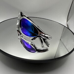 Op de afbeelding: Chromen zonnebril met blauw-paarse verlopende glazen. De zonnebril wordt weerspiegeld in een ronde spiegel. Het montuur heeft een uniek, hoekig ontwerp. De glazen lopen over van blauw naar paars.