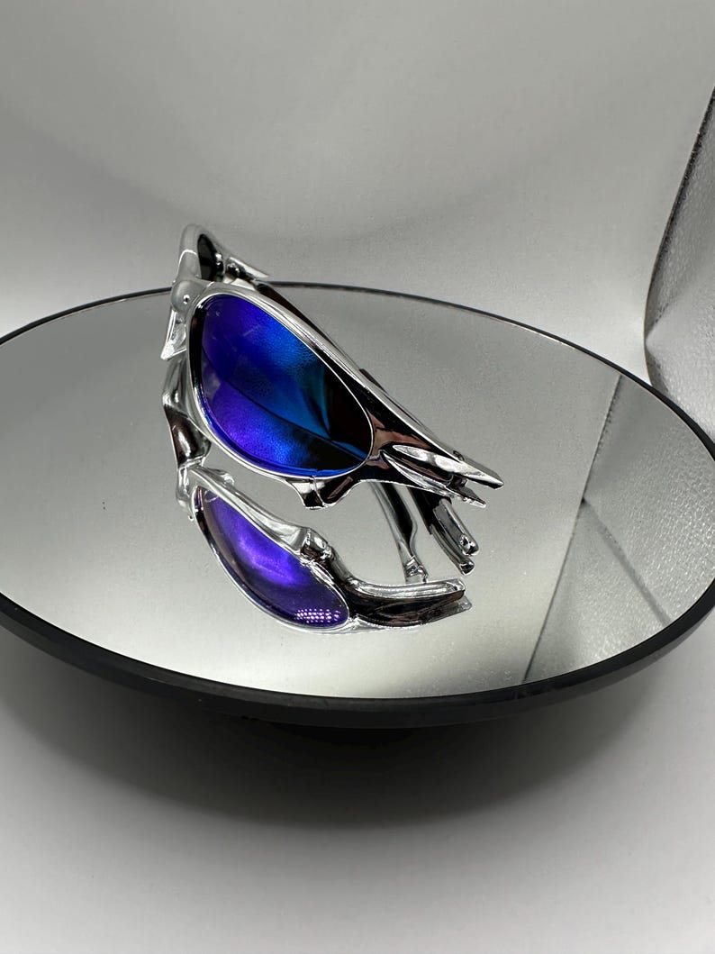 Op de afbeelding: Chromen zonnebril met blauw-paarse verlopende glazen. De zonnebril wordt weerspiegeld in een ronde spiegel. Het montuur heeft een uniek, hoekig ontwerp. De glazen lopen over van blauw naar paars.