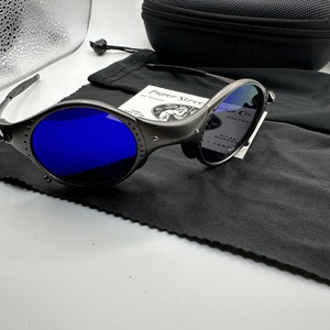 May include: A pair of blue mirrored sunglasses with a silver frame and a black carrying case. The sunglasses have a unique design with a small round lens on the side. The case is made of a soft, black material and has a zipper closure.