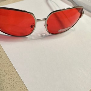 Puede incluir: Gafas de sol con montura plateada y lentes rojos. Las gafas est&aacute;n sobre una superficie blanca.