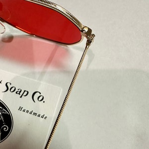 Może przedstawiać: Okulary przeciwsłoneczne z metalu w kolorze złotym z czerwonymi szkłami. Okulary są złożone na p&oacute;ł, a zausznik leży na białej kartce z tekstem "Soap Co. Handmade Tyler Durden 1980".