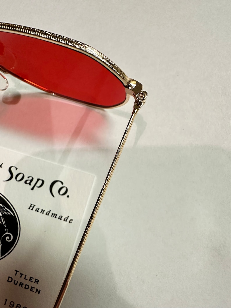 Może przedstawiać: Okulary przeciwsłoneczne z metalu w kolorze złotym z czerwonymi szkłami. Okulary są złożone na p&oacute;ł, a zausznik leży na białej kartce z tekstem "Soap Co. Handmade Tyler Durden 1980".