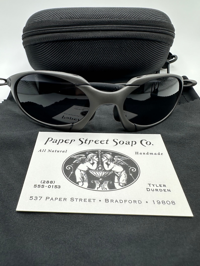 May include: A gray and black pair of sunglasses with a black case. The sunglasses are on a gray fabric surface with a white business card that reads "Paper Street Soap Co.", "All Natural", "Handmade", "(288) 555-0153", "537 Paper Street. Bradford. 19808", "Tyler Durden".