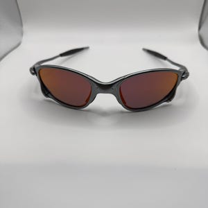 May include: A pair of sunglasses with a dark gray frame and red-tinted lenses. The sunglasses have a sleek, modern design, with a curved frame and slender arms. The lenses are a vibrant red color, providing a striking contrast to the frame.