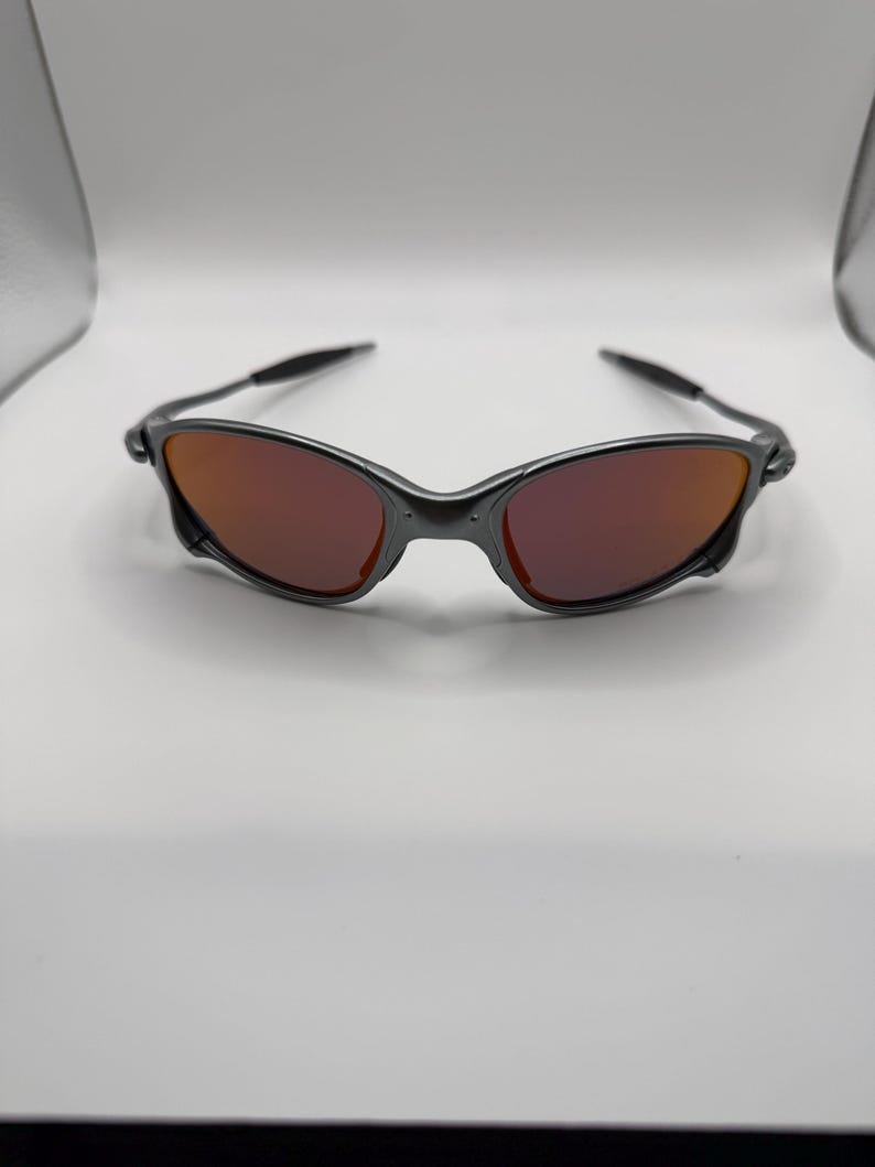 May include: A pair of sunglasses with a dark gray frame and red-tinted lenses. The sunglasses have a sleek, modern design, with a curved frame and slender arms. The lenses are a vibrant red color, providing a striking contrast to the frame.